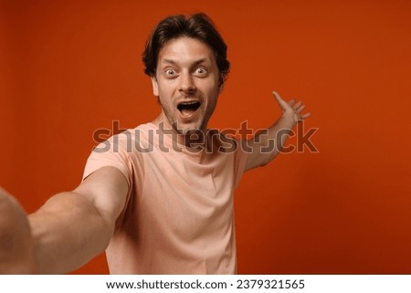 Excited young man wearing t-shirt taking selfie and gesturing while standing isolated over red studio background