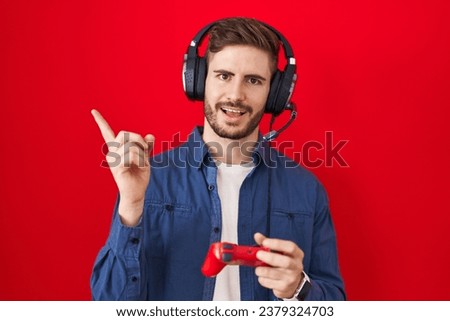 Hispanic man with beard playing video game holding controller smiling happy pointing with hand and finger to the side 