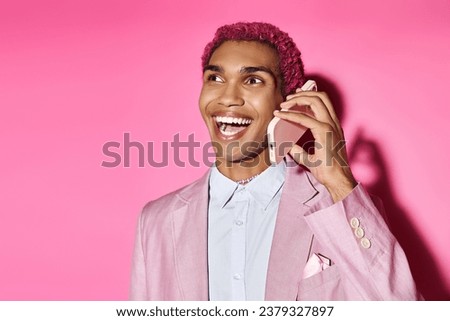 cheerful young african american male model in pink blazer posing with mobile phone on pink backdrop