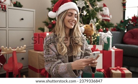Young blonde woman using smartphone sitting by christmas tree at home Royalty-Free Stock Photo #2379336847