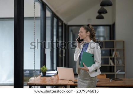 Smart businesswoman stands holding documents and talking on the phone at the table in the office.