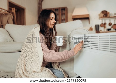 Cold home, freezing. Using heater at home in winter. Woman warming her hands sitting by device and wearing warm clothes and holding hot drink. Heating season.  Royalty-Free Stock Photo #2379350095