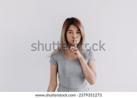 Asian Thai woman wear grey, hold fingers on lips, showing shh, hush quiet sign, making gesture silence, keep secret, looking at camera, isolated on white background wall.