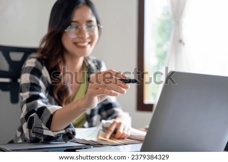 Young female architect or interior designer sits at her desk in the office, she points at laptop screen and selects colors from charts, envisioning the entire project. 