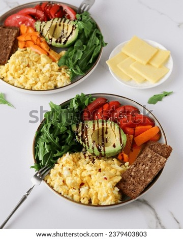 Elegant breakfast plate: creamy scrambled eggs paired with ripe avocado, fresh tomatoes, arugula, and bell pepper, enhanced by balsamic drizzle, sesame seeds, and chili flakes for a spicy kick