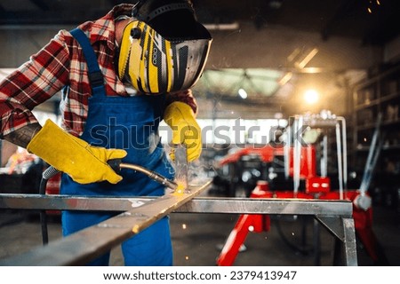 Worker in a dangerous work environment welding metal while wearing a safety equipment and looking down at the flying sparks. Blurred background of a working space and shelves with tools.Copy space.