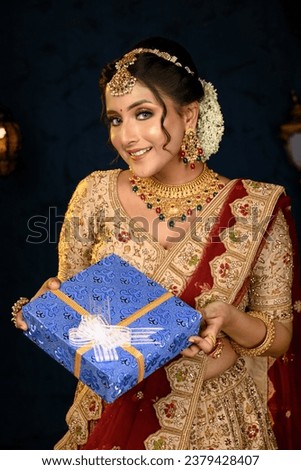 Portrait of a pretty young Indian woman dressed in traditional lehenga, gold jewellery and bangles holding gift box in hands on decorative background. Indian culture, occasion, religion and fashion.