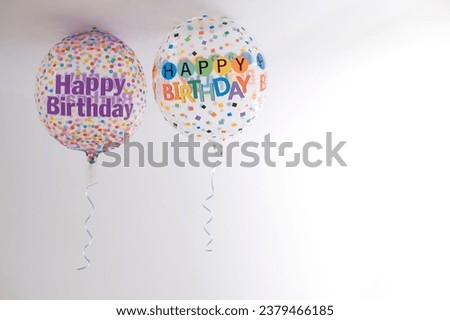two helium balloons decorated with colored dots and a happy birthday message, with a hanging ribbon, on the roof of a white room, copy space, degraded background, horizontal