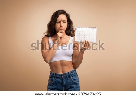 Confused upset brunette millennial woman with calendar touching her chin, isolated on beige studio background. Early menopause, late period, unintended pregnancy