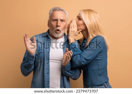 Attractive mature blonde woman sharing secret or whispering gossips into her husband ear, beige studio background. Loving elderly spouses sharing rumors. Communication in marriage Royalty-Free Stock Photo #2379473707