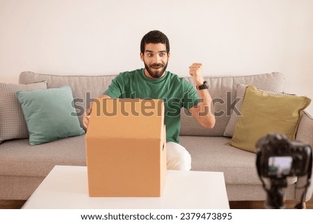 Satisfied millennial arab man blogger unpacks cardboard box, celebrate win, enjoy success sign, making video review on camera in home office interior. Delivery and blog, online shopping gesture