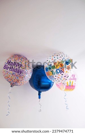 four helium balloons decorated with colored dots and a happy birthday message, with a hanging ribbon, on the roof of a white room, degraded background, copy space, vertical