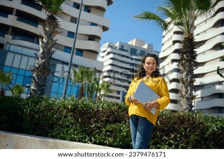 Bottom view of a charming Latin American curly freelance woman, entrepreneur holding a laptop, smiling looking aside, standing against high-rise modern office buildings background on a warm sunny day