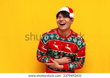 young asian man in christmas sweater and santa hat laughs and makes fun of on yellow isolated background, korean guy in new year clothes jokes and shows ridicule