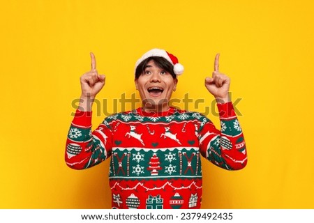 young asian man in christmas sweater and santa hat pointing with hands up at copy space on yellow isolated background, korean guy in new year clothes showing and advertising empty space on top