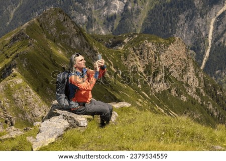 Gray-haired man with backpack sitting on rocky ledge and taking pictures of summer mountain landscape, Austria