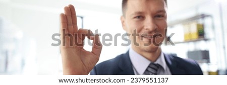 Smiling businessman shows OK sign. Cheerful office worker performs tasks successfully. Professional manages affairs on blurred background
