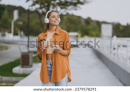 Young minded fun woman wear orange shirt casual clothes listen music in headphones use mobile cell phone drink coffee rest relax walk in spring city park outside in summer day. Urban lifestyle concept