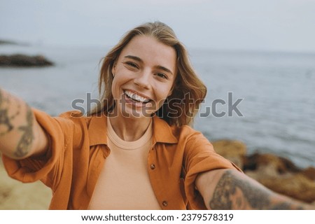 Close up young smiling happy woman she wears orange shirt casual clothes doing selfie shot pov on mobile cell phone walk on sea ocean sand shore beach outdoor seaside in summer day. Lifestyle concept