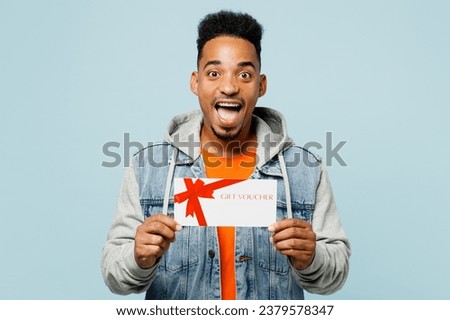 Young fun man of African American ethnicity wear denim jacket orange t-shirt hold gift certificate coupon voucher card for store isolated on plain pastel light blue cyan background. Lifestyle concept