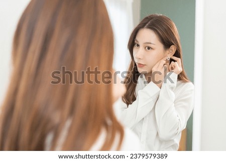 Get dress, pretty asian young woman, businesswoman standing putting earring or jewelry, wearing white shirt formal getting dressed getting ready before go to work looking reflection the mirror at home