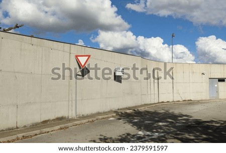 Yield sign on sidewalk by side of a long surrounding concrete wall. Street in front, blue cloudy sky on behind. Background for copy space