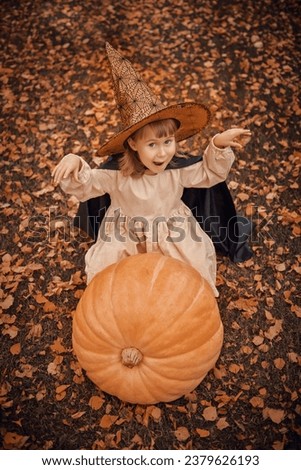 Caroling for Halloween. A cute little girl, dressed up in a witch costume, stands on a large pumpkin with a basket of sweets and smiles.