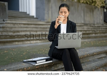 A busy, professional, and concentrated millennial Asian businesswoman in a formal business suit is sipping coffee and reading an urgent email on her laptop while sitting on the stairs in the city.