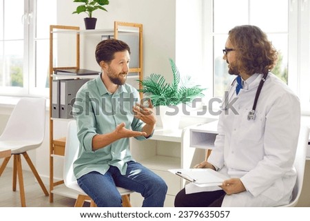 Portrait of bearded young man visiting male doctor for medical examination. Sick patient listening to doctor consultation in clinic during checkup in exam room. Healthcare and medicine concept. Royalty-Free Stock Photo #2379635057