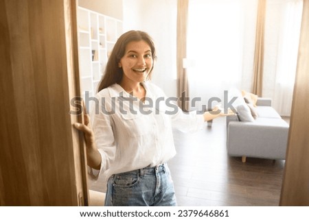 Cheerful young Indian woman with welcoming gesture, standing by open door, expressing joy and warmth in homey and well-lit living room setting, free space Royalty-Free Stock Photo #2379646861