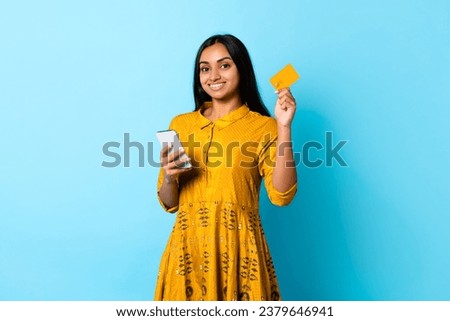 Smiling young Hindu lady in yellow dress using debit card and smartphone for online shopping on blue studio background. Embracing modern ecommerce and banking applications