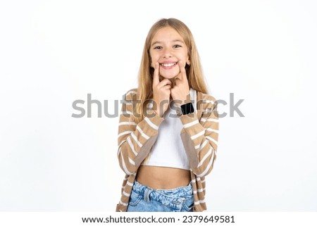Happy beautiful caucasian kid girl with toothy smile, keeps index fingers near mouth, fingers pointing and forcing cheerful smile