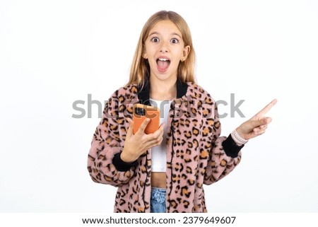 Astonished beautiful kid girl wearing animal print jacket holding her telephone and pointing with finger aside at empty copy space