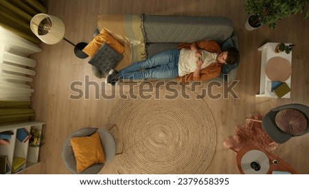Top view capturing a young man laying on the couch, looking at the screen of his smartphone. He is texting, chatting with someone online. Royalty-Free Stock Photo #2379658395