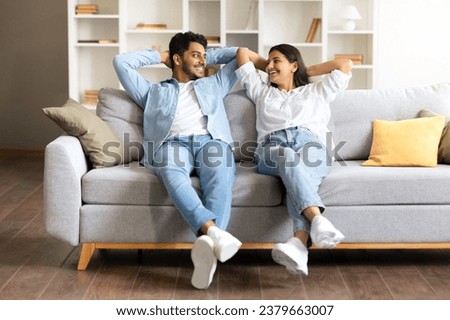 Indian young couple dressed casually, share light-hearted moment on comfortable sofa in homey living room with soft pillows and neat bookshelves in the background