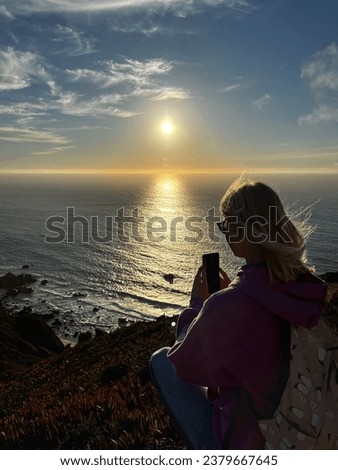 Woman in violet hoodie and sunglasses making photo of sunset on the shore of the Atlantic ocean using mobile phone