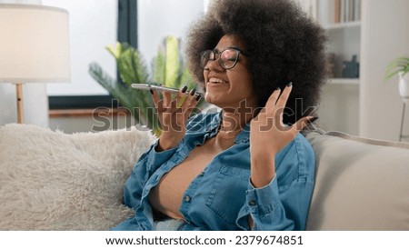 Ethnic girl African American woman talk virtual assistant on mobile phone smiling lady recording answering audio message voice mail in social network for friend using loudspeaker talking at home sofa