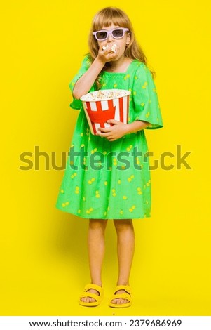 Excited young cute school girl in 3D glasses eating popcorn, watching interesting tv serial, sport game, film, online social media movie content. Teen female child kid on yellow background. Vertical