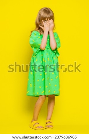 Nosy curious blonde child girl kid closing eyes with hands, spying through fingers, hiding peeping, exploring way, seeking something in distance. Little preteen children on yellow background. Vertical Royalty-Free Stock Photo #2379686985