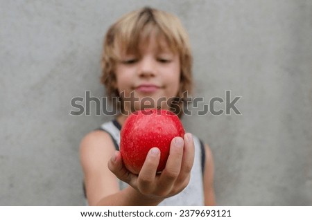 Handsome out of focus 6 year old caucasian blond kid holding and showing a red apple with vitamins looking happy outside. Isolated concrete background. Concept of healthy eating in childhood