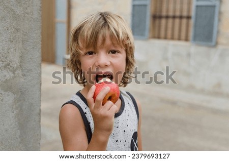 Smiling happy six year old caucasian blond boy eating open mouth a red apple with vitamins outside of the house. Concept of healthy lifestyle and food habits in childhood.  Horizontal photography