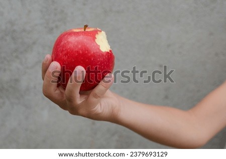 Child hand holding bitten red apple with vitamins isolated on concrete background. Horizontal photograph. Concept of healthy lifestyle and good eating habits in childhood
