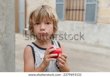Cute six seven year old caucasian blond boy happily eating red apple with vitamins looking at camera outside of the house. Concept of healthy lifestyle and food habits in childhood. Horizontal image