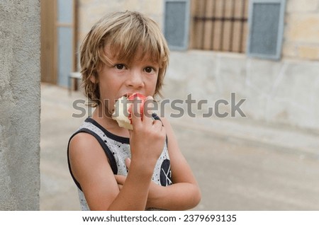 Adorable six years old caucasian blond boy eating confident a red apple with vitamins outside of the house. Concept of healthy lifestyle and food habits in childhood.  Horizontal photography