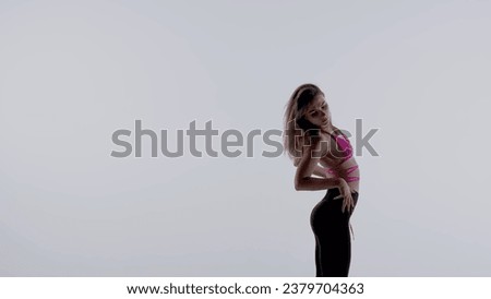 Young woman wearing a bright pink bra, top and leggings performing energetic heels dance or posing in studio, isolated white background. Medium full.