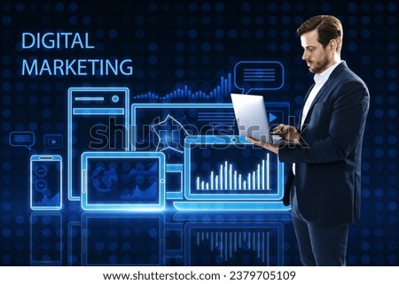 Attractive young european male using laptop while standing on blue pixel background with gadgets and business chart hologram. Digital marketing, finance, social network and online service concept