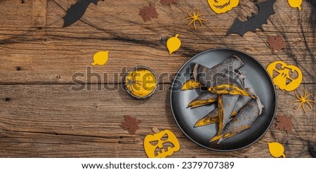 Halloween sweet dessert with traditional decor. Thin black pancakes with pumpkin cream, fall leaves, spiders and bats. Spider web, wooden background, hard light, dark shadow, banner format