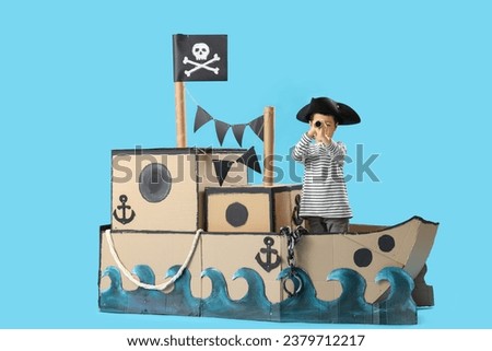 Cute little pirate with spyglass in cardboard ship on blue background Royalty-Free Stock Photo #2379712217