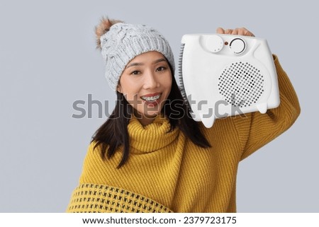 Young Asian woman in winter clothes with electric heater near light wall