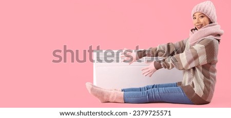 Young Asian woman and electric heater on pink background with space for text. Heating season